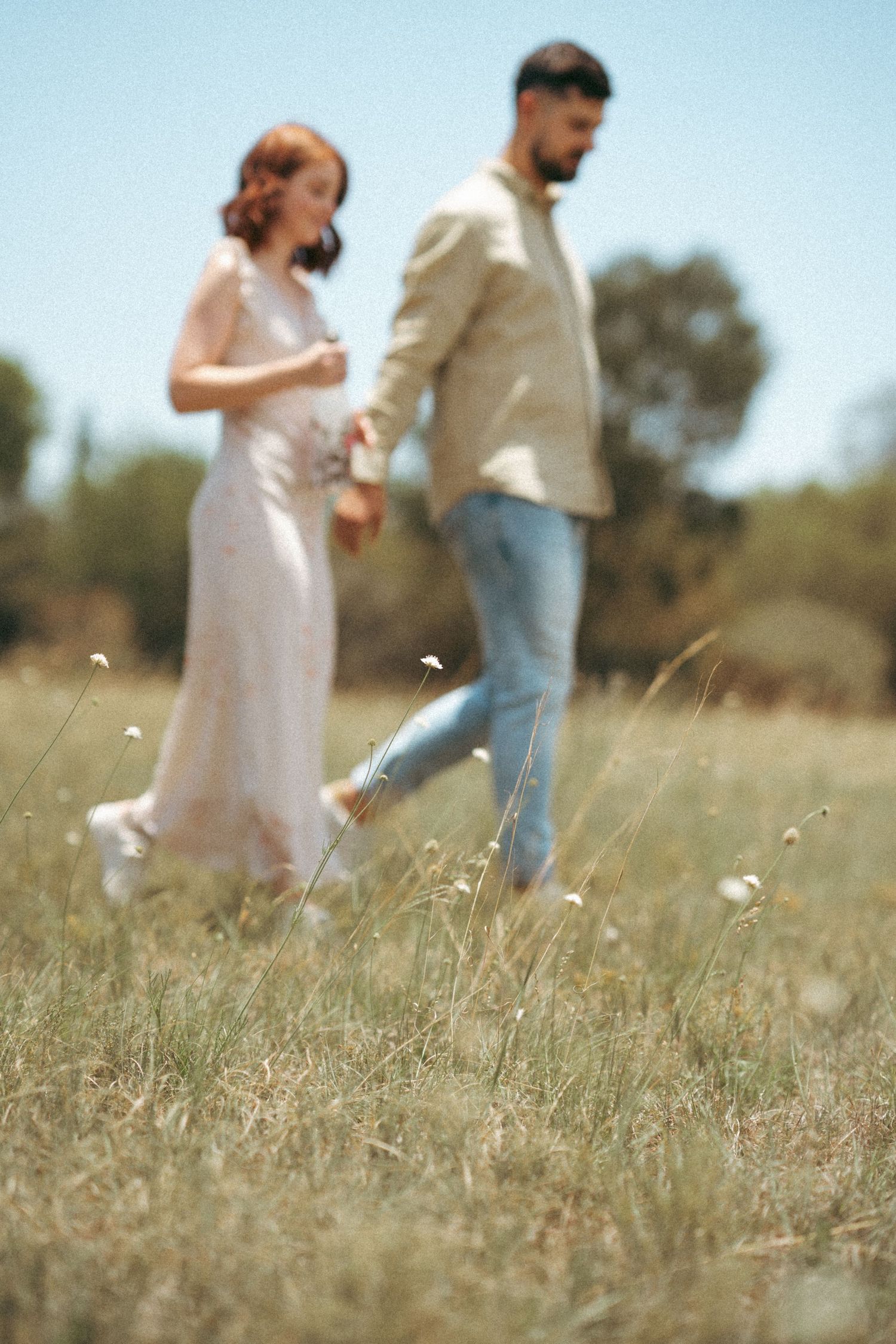 A couple walks together through a sunlit field of tall grass on a warm summer day.