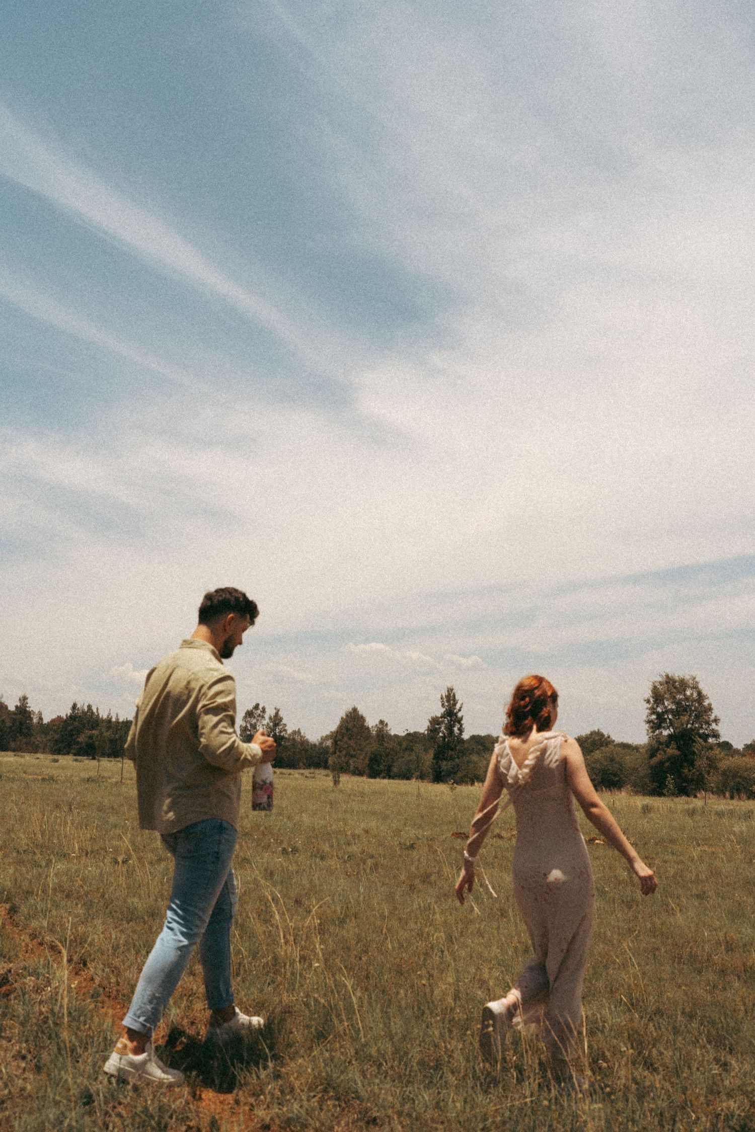 Two people chase each other through a golden field under wispy clouds.