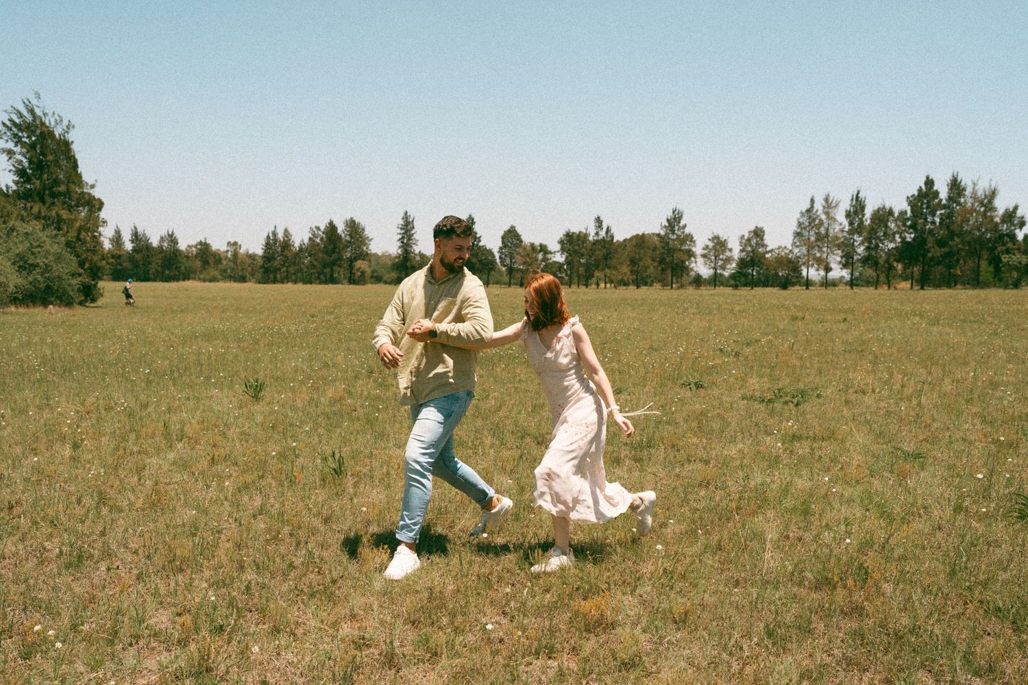 A couple dances playfully in a sunlit field with trees in the background.
