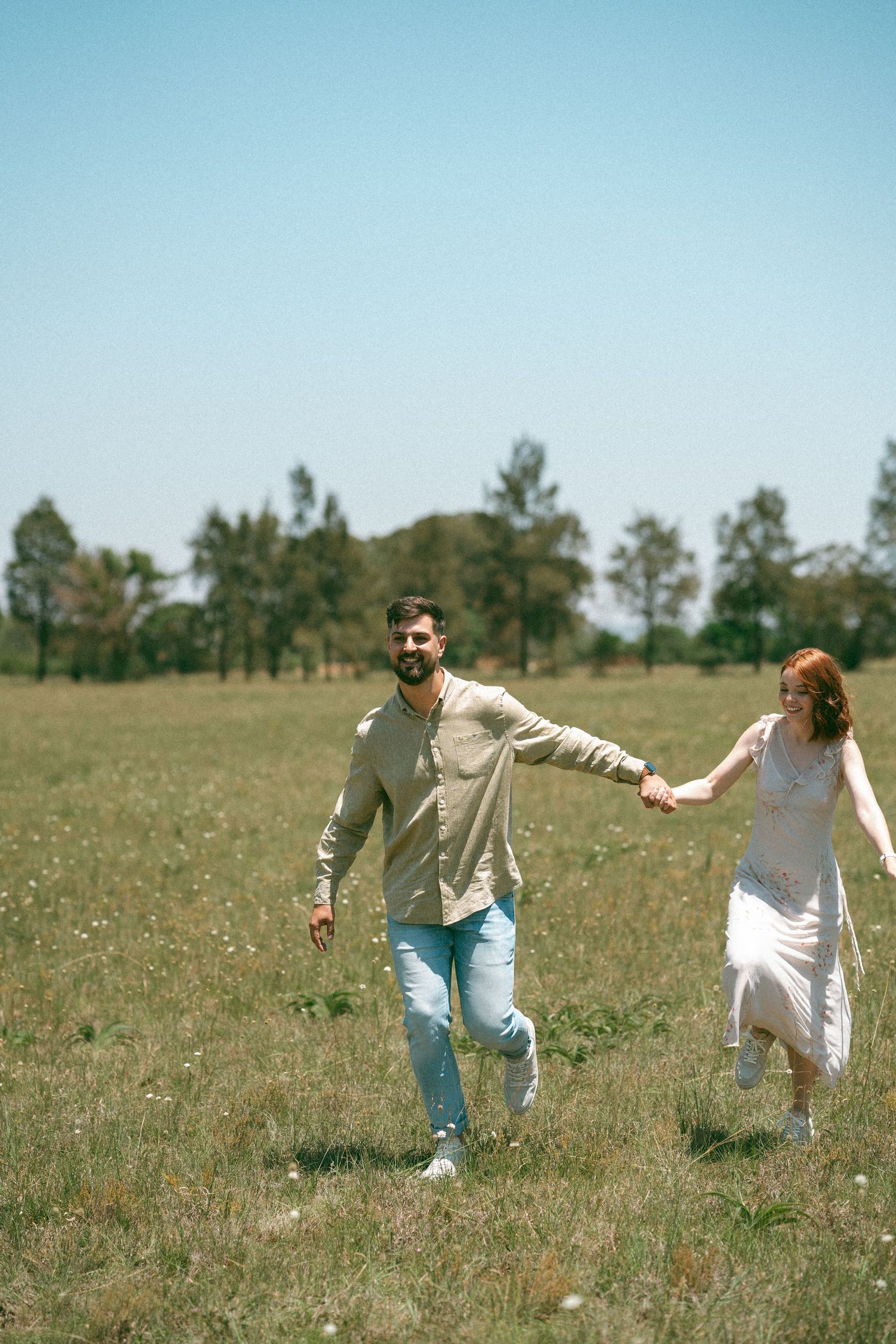 A couple playfully runs and dances together in a grassy meadow on a sunny day.