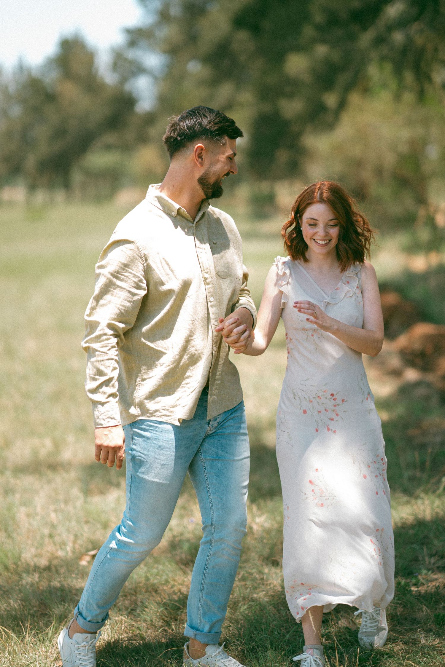 A couple walks together through a sunny field, sharing joyful laughter in casual summer attire.