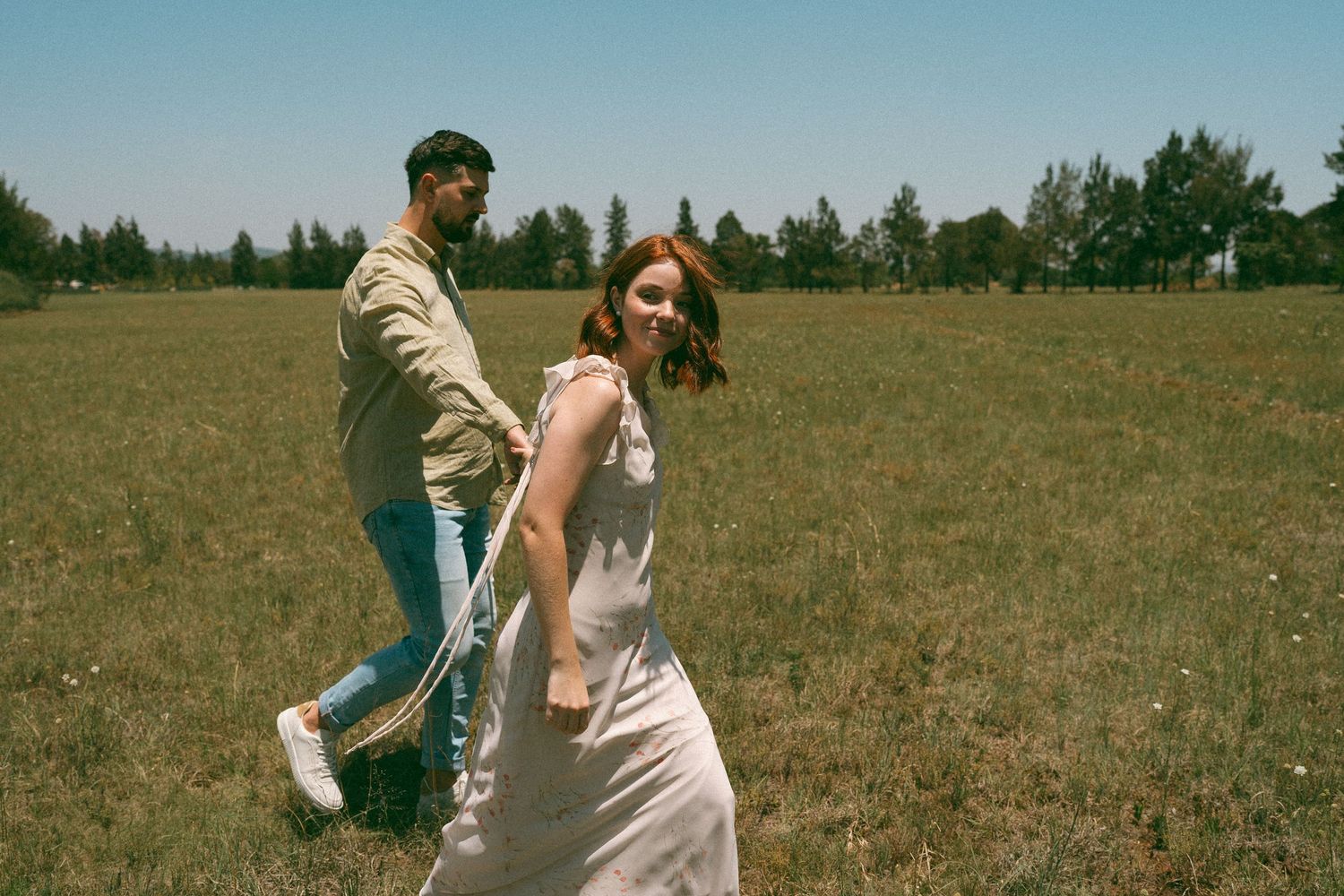 A couple dances carefree in a grassy field with evergreen trees in the background.