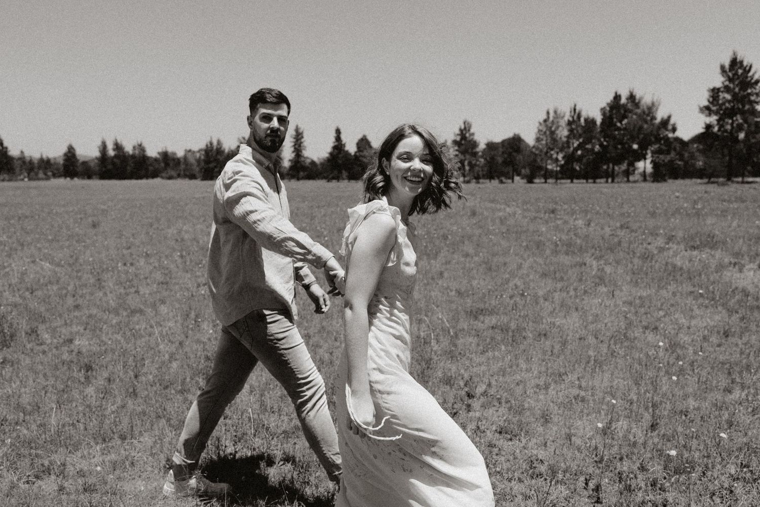 A black and white photo captures a couple dancing playfully in a meadow.