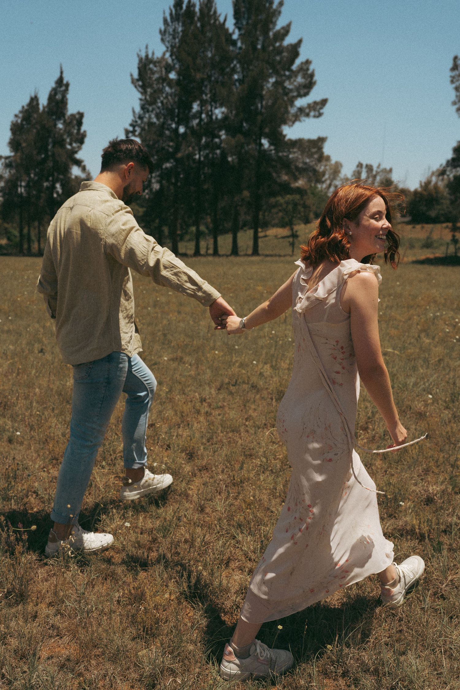 A couple holds hands while skipping through a sunlit field near pine trees.