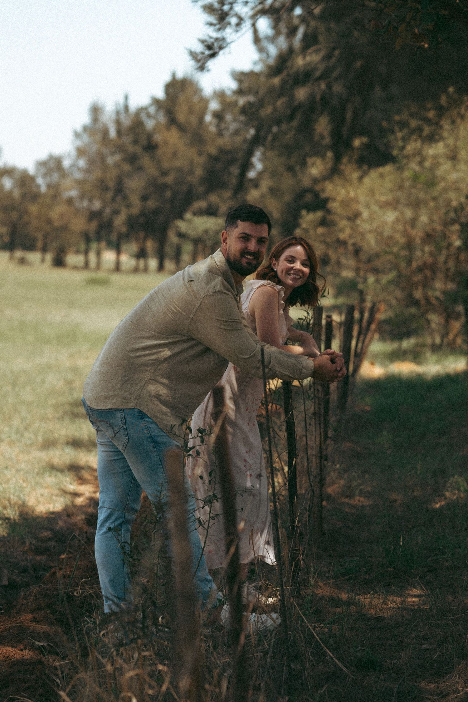 A couple shares a tender moment while leaning against a wooden fence in a pastoral setting.