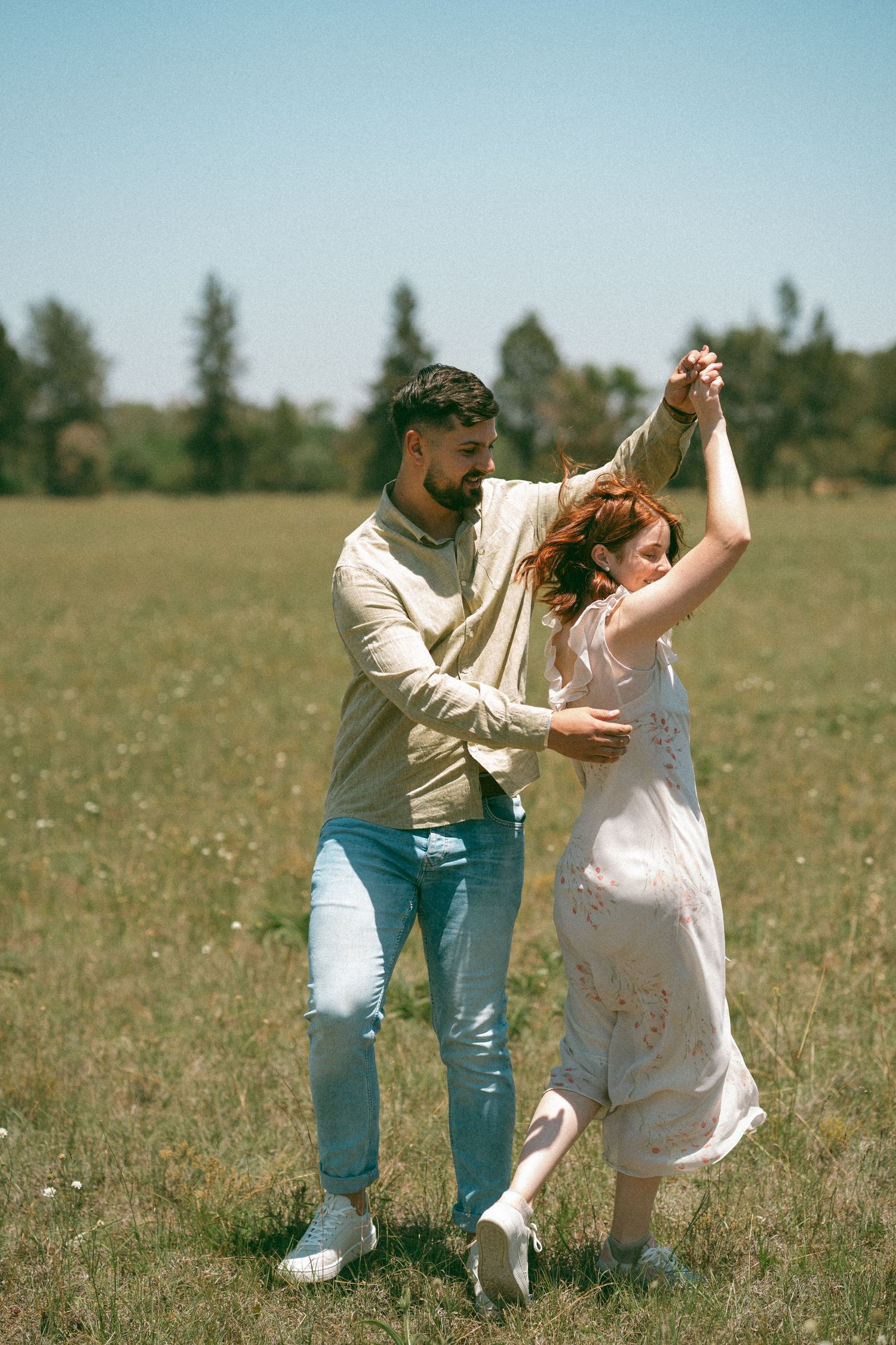 A couple dances playfully in a sunny meadow wearing casual clothing and enjoying a carefree moment together.