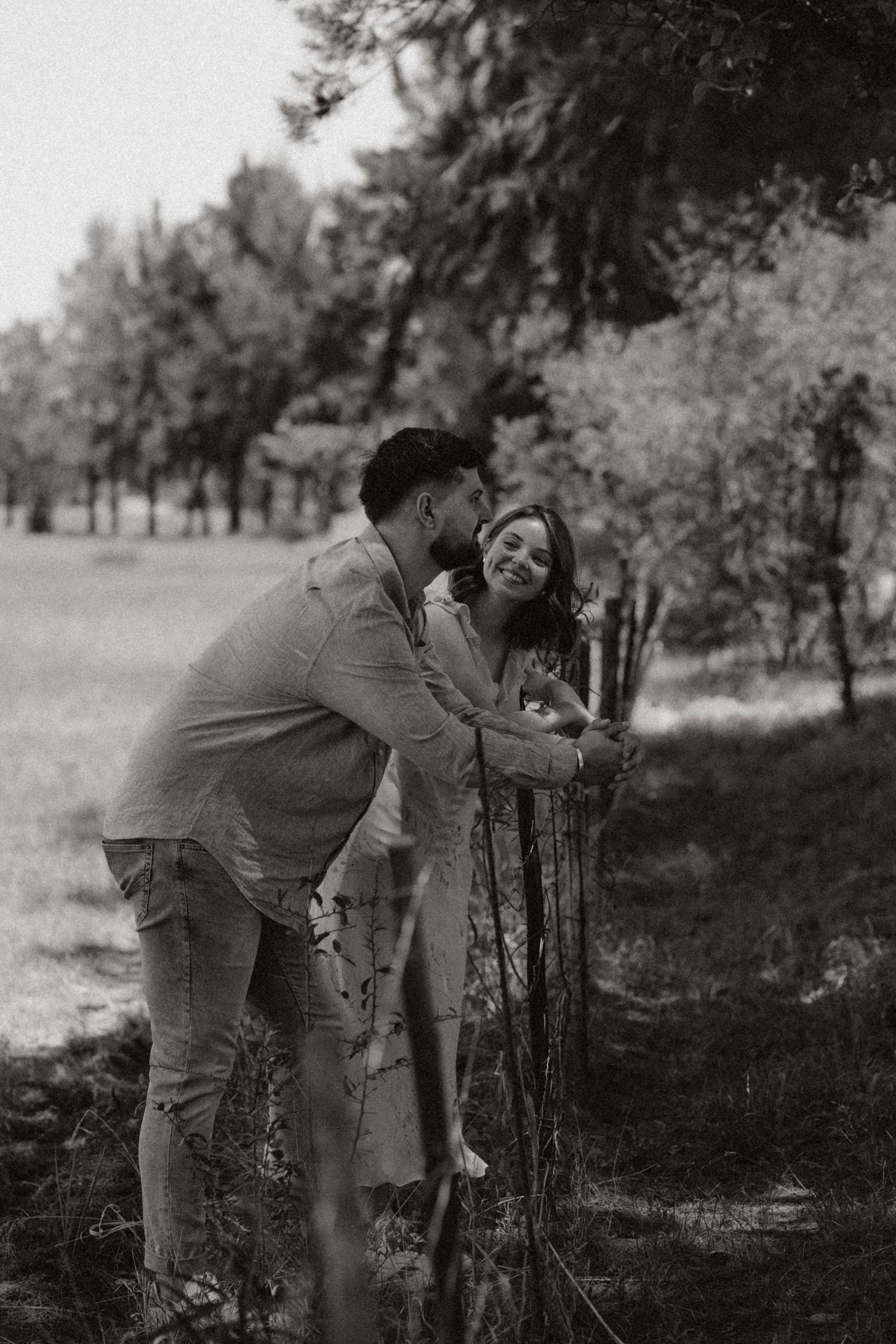 A black and white image shows a couple sharing a laugh while leaning against a rustic fence.