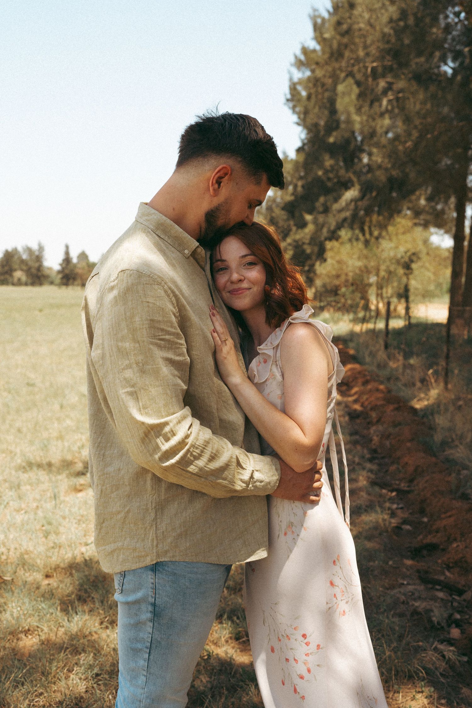 A tender embrace between a couple standing in a field with trees in the background.