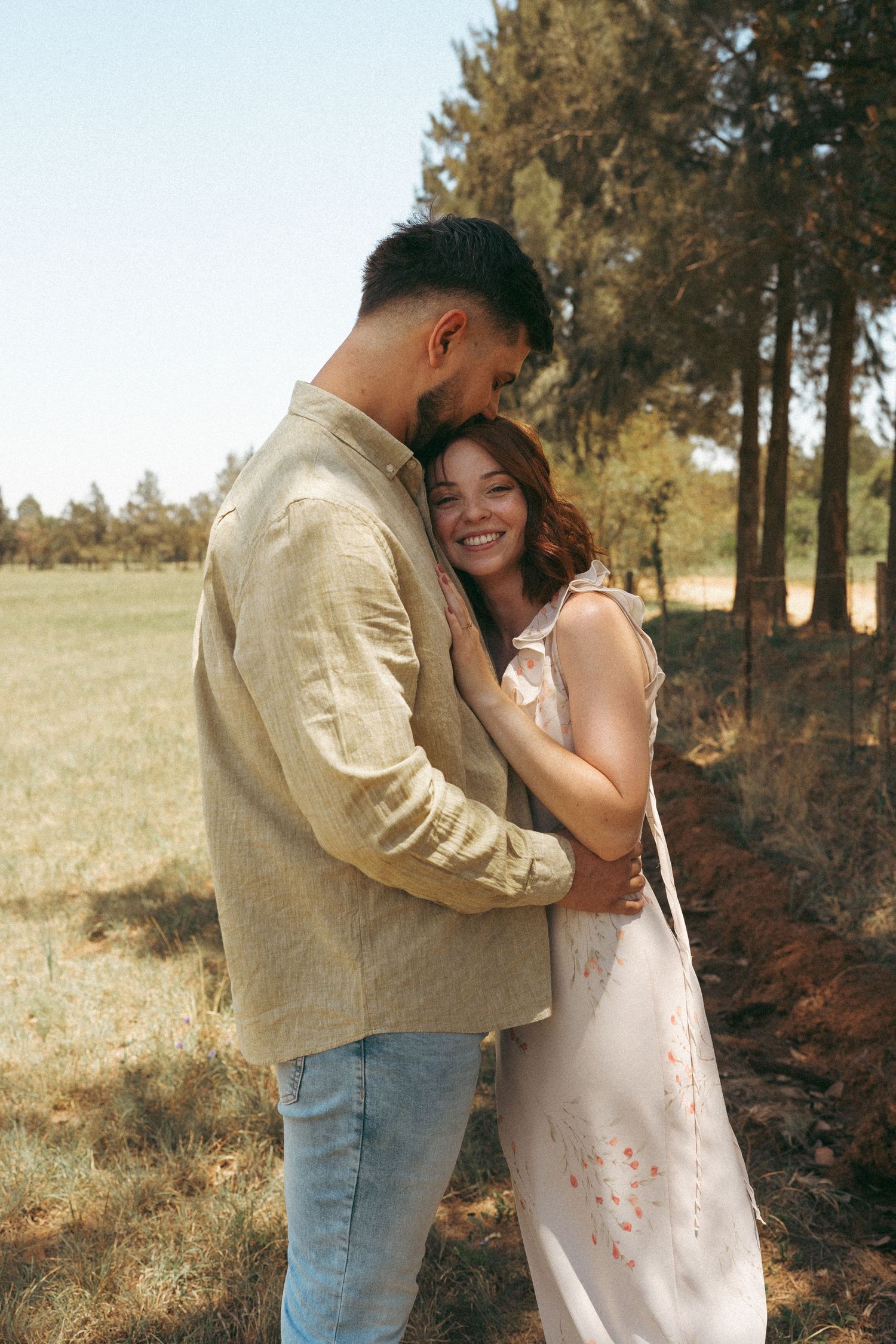Romantic couple embraces in a sunlit field with pine trees in the background.