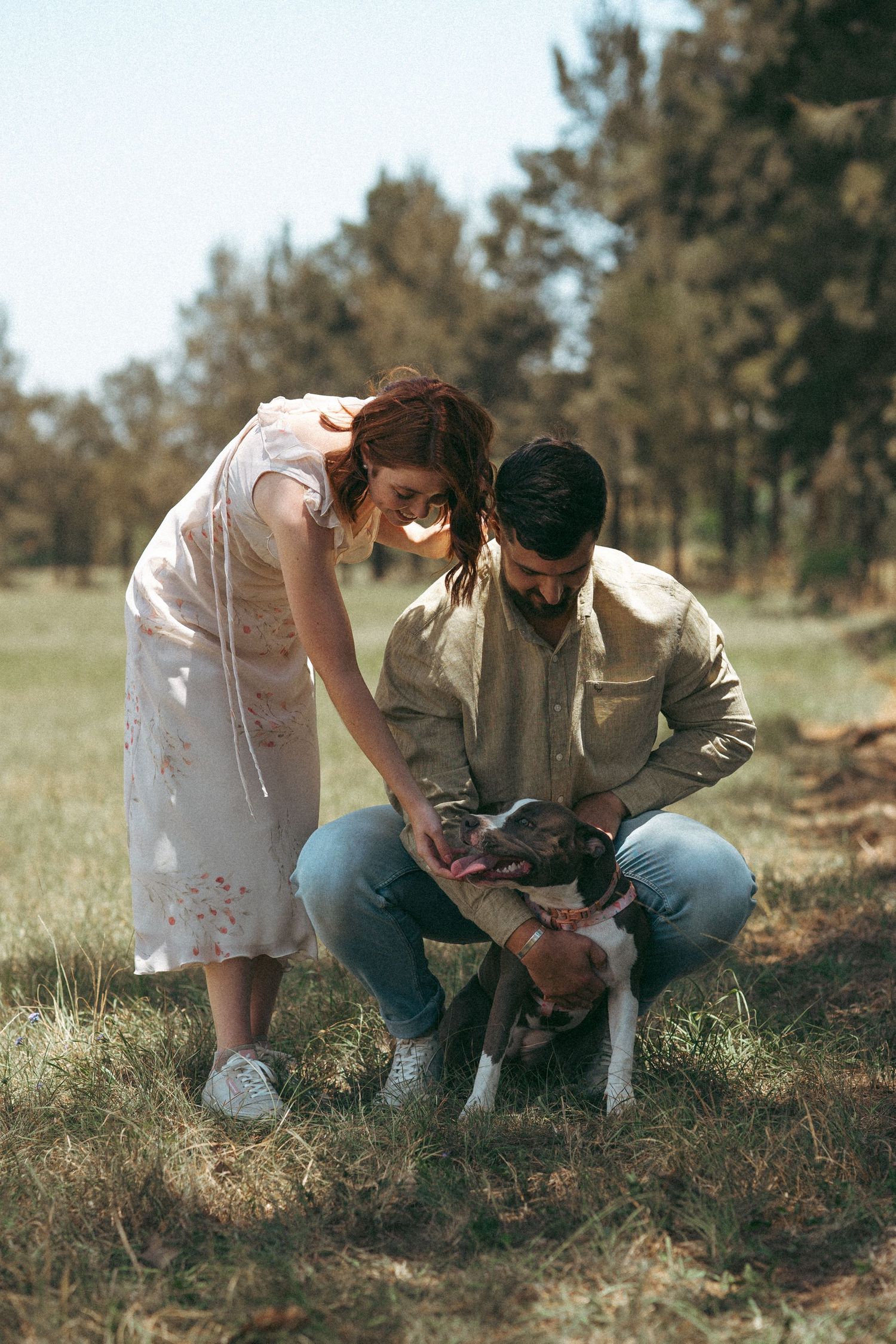 Couple crouches together examining something in the grass outdoors.