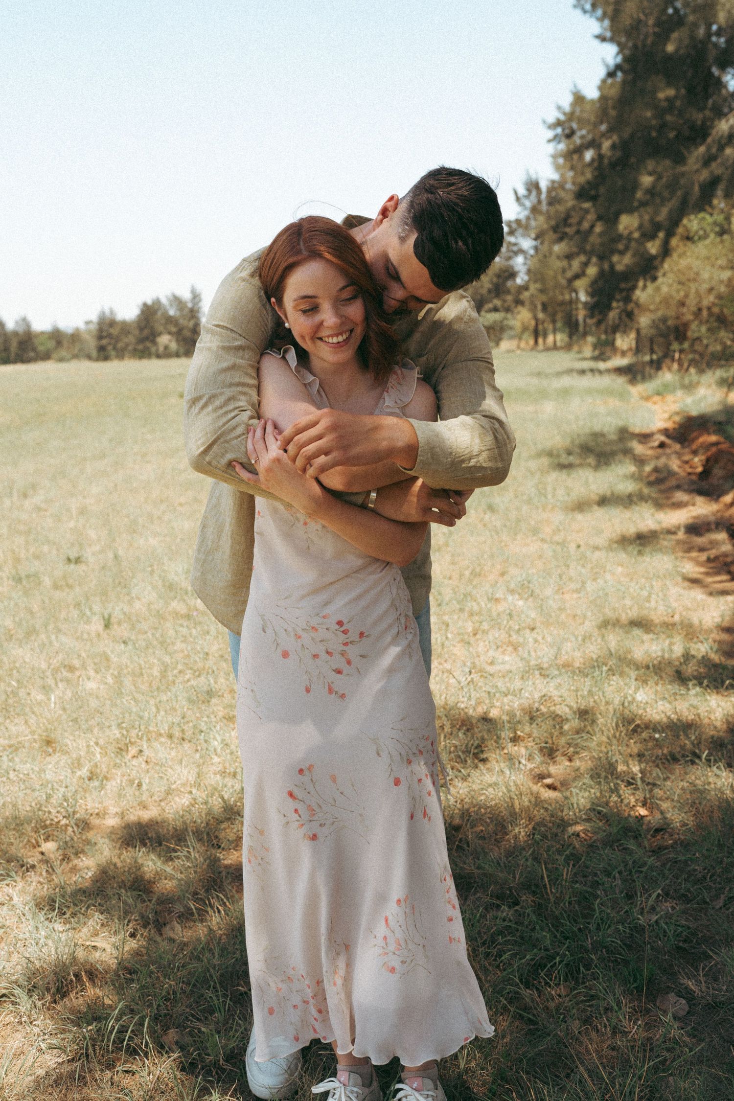 Couple embracing in a sunlit meadow wearing light colored clothing.
