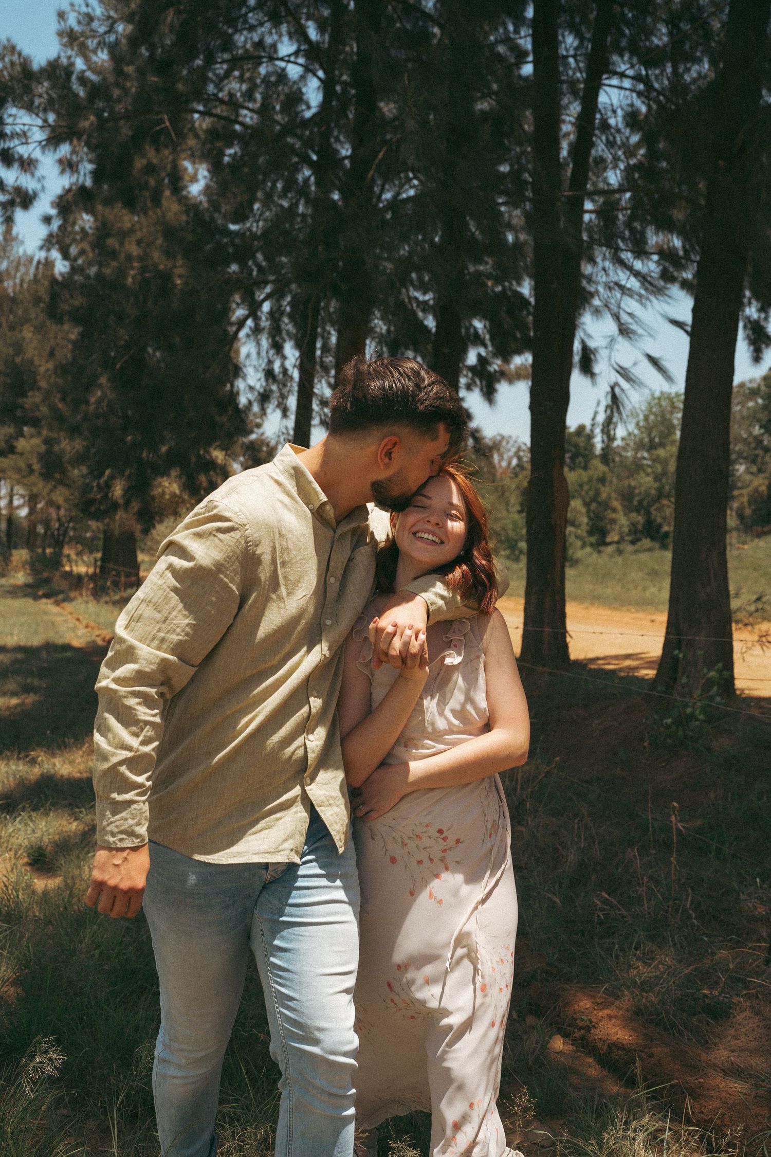 A couple shares playful moments together among pine trees on a sunny afternoon.