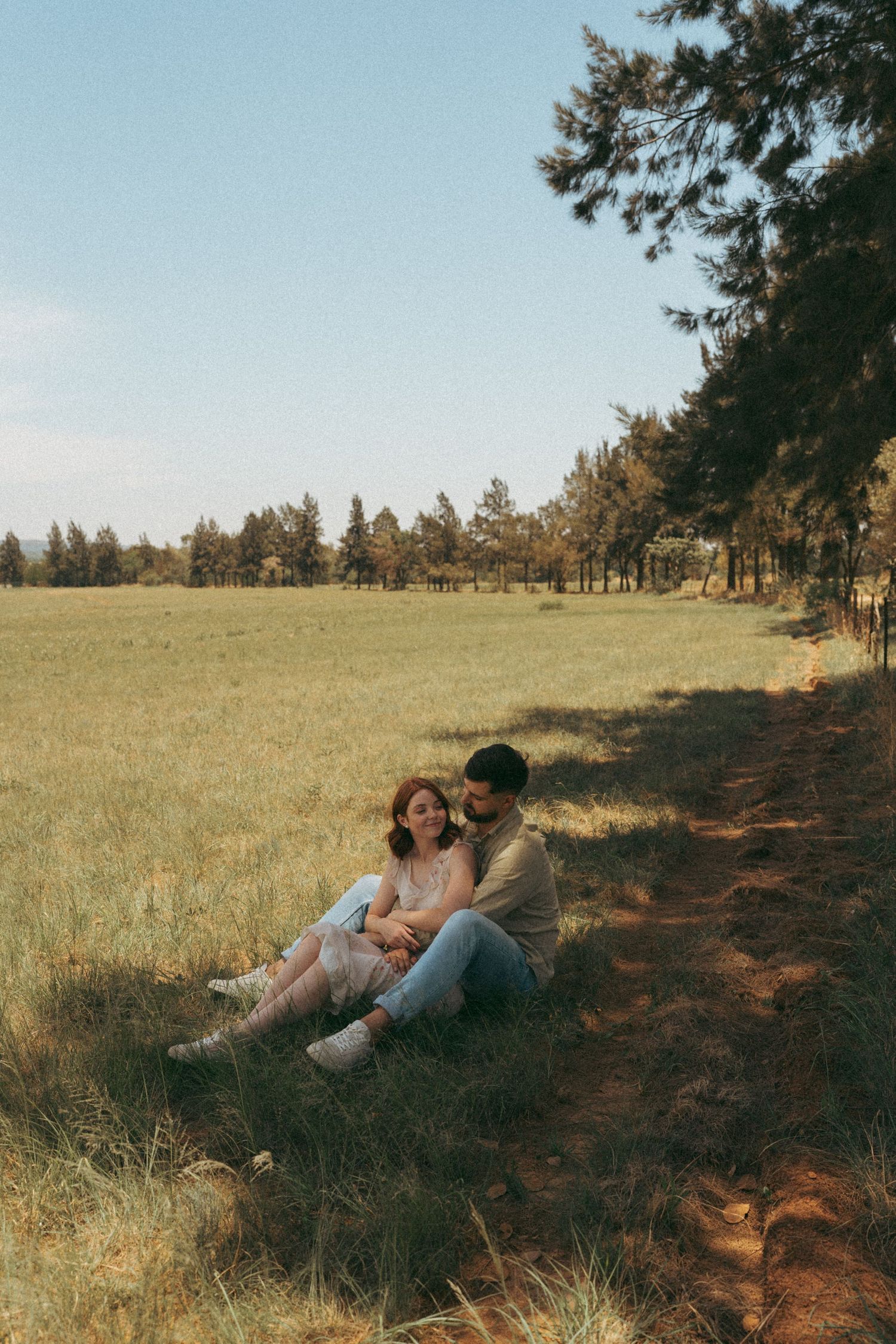 Couple lying together on grassy hillside with pine trees in background during golden hour.