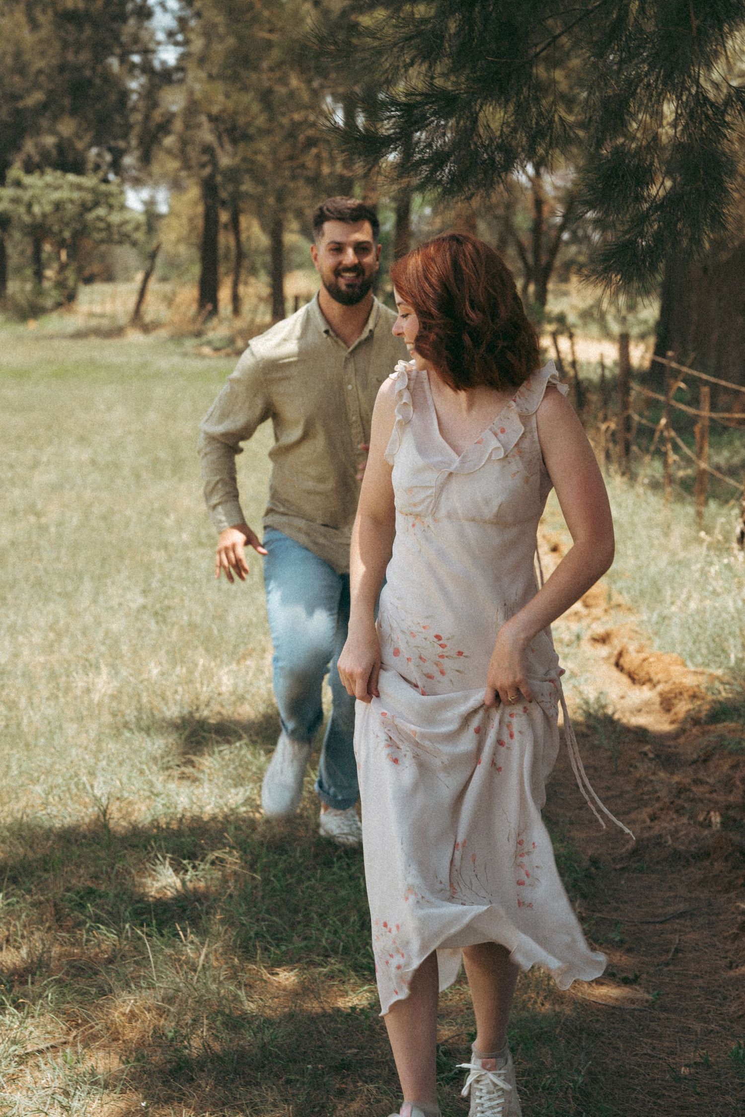 A playful couple walking along a wooded trail, with her wearing a flowing white dress.