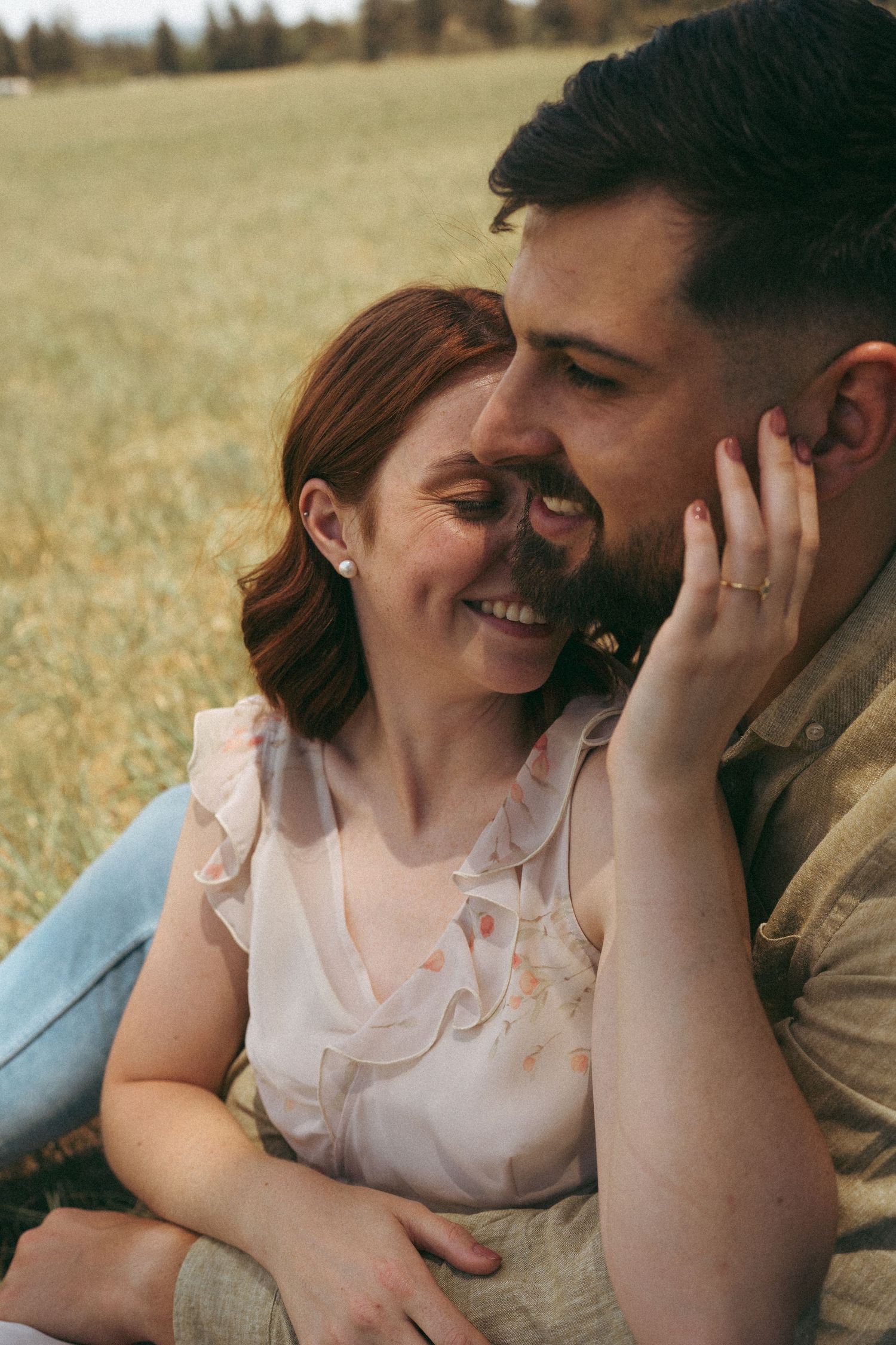 A romantic couple shares an intimate moment in a sunlit meadow, with her hand gently touching his face.
