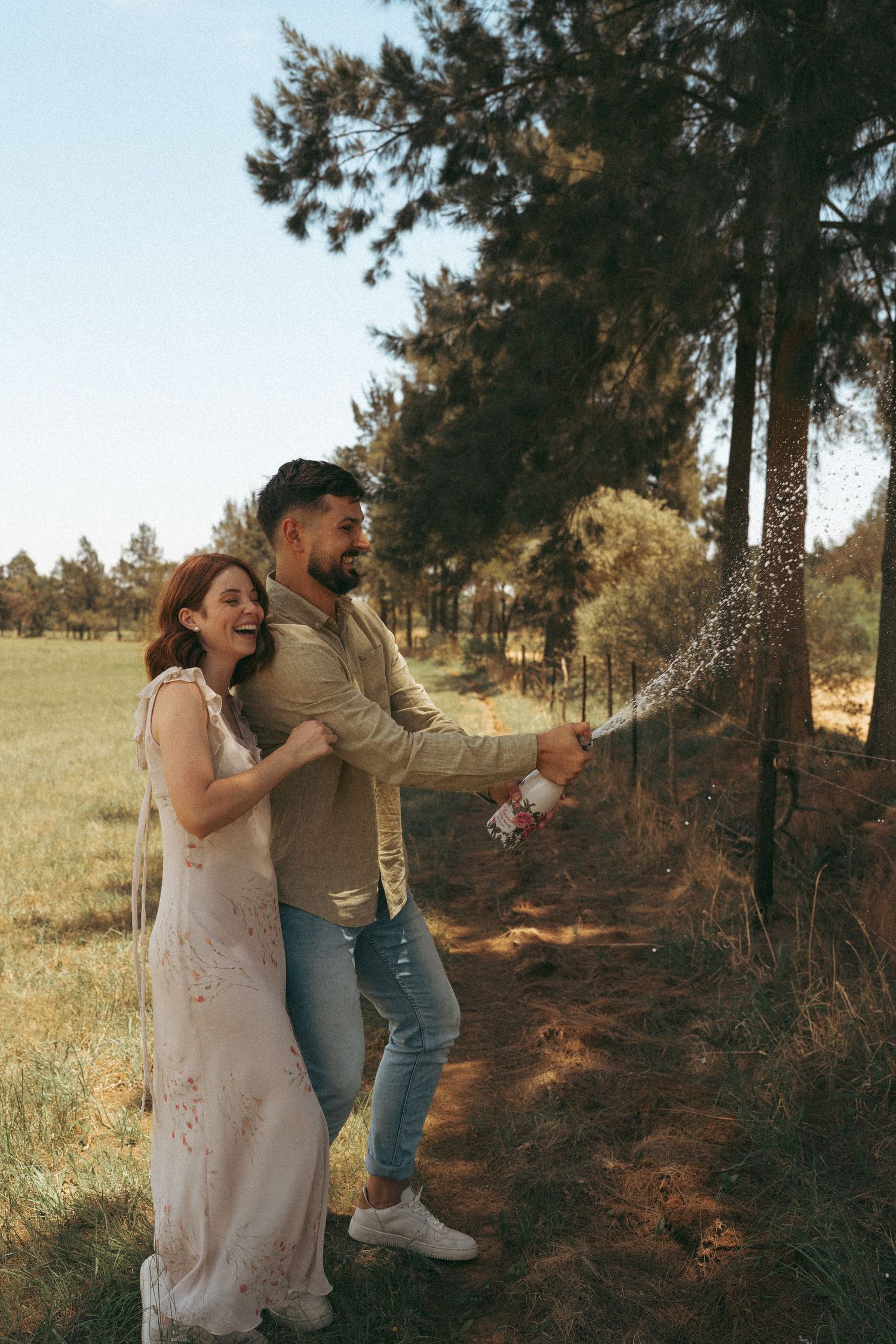 A couple sprays champagne while celebrating outdoors near pine trees.