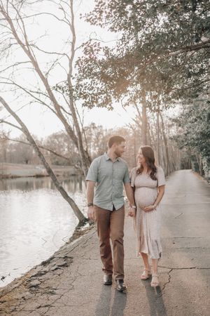 A couple walks along a lakeside path during sunset with trees casting dappled light on the peaceful scene.