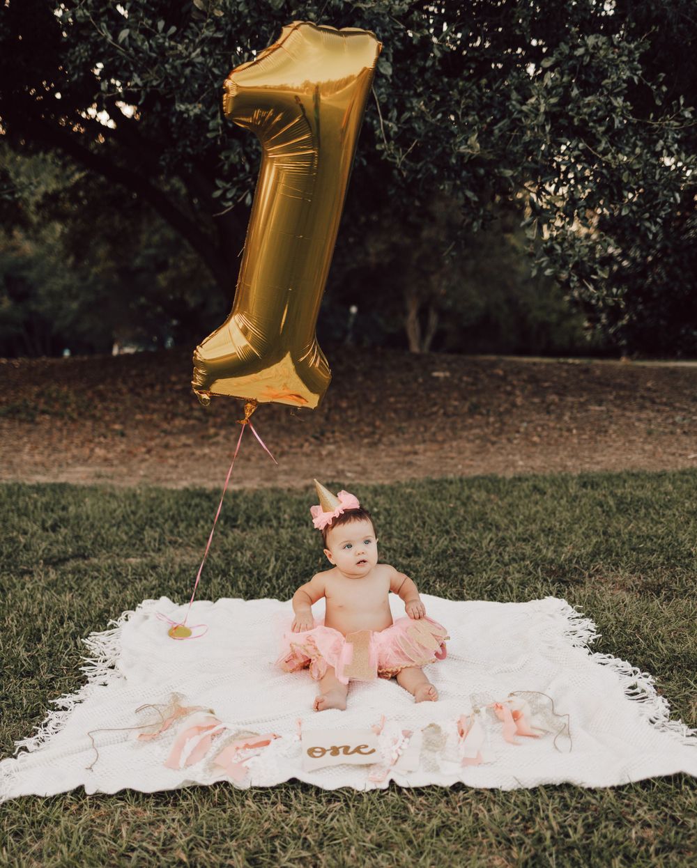 Baby celebrates first birthday with gold balloon and pink decorations on white blanket.