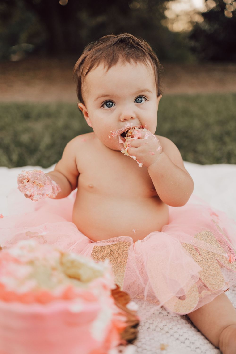 Baby wearing pink tutu messily eating birthday cake at outdoor celebration.