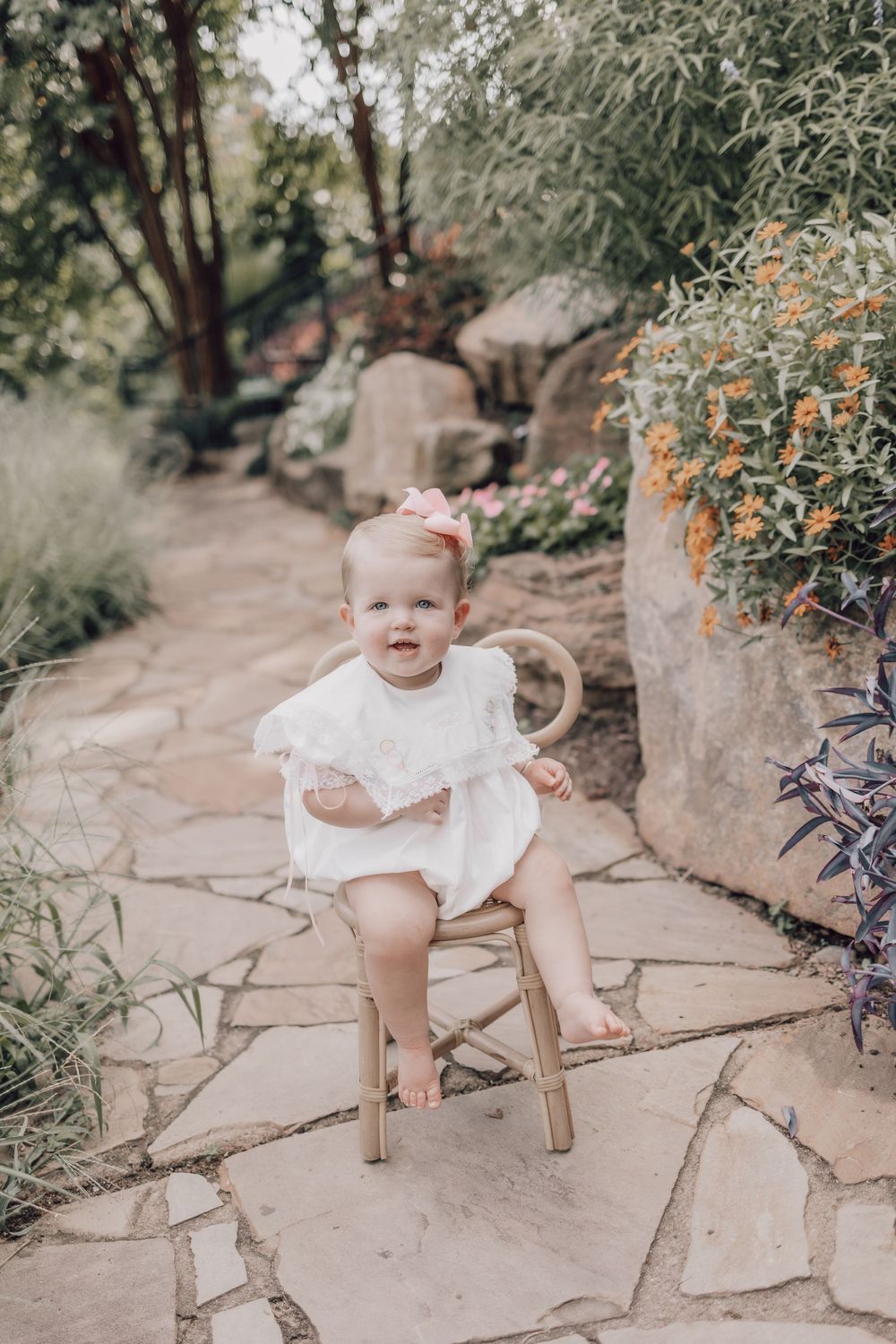 In a garden setting with stone paths and orange flowers, a baby sits in a small wooden chair wearing a white dress.