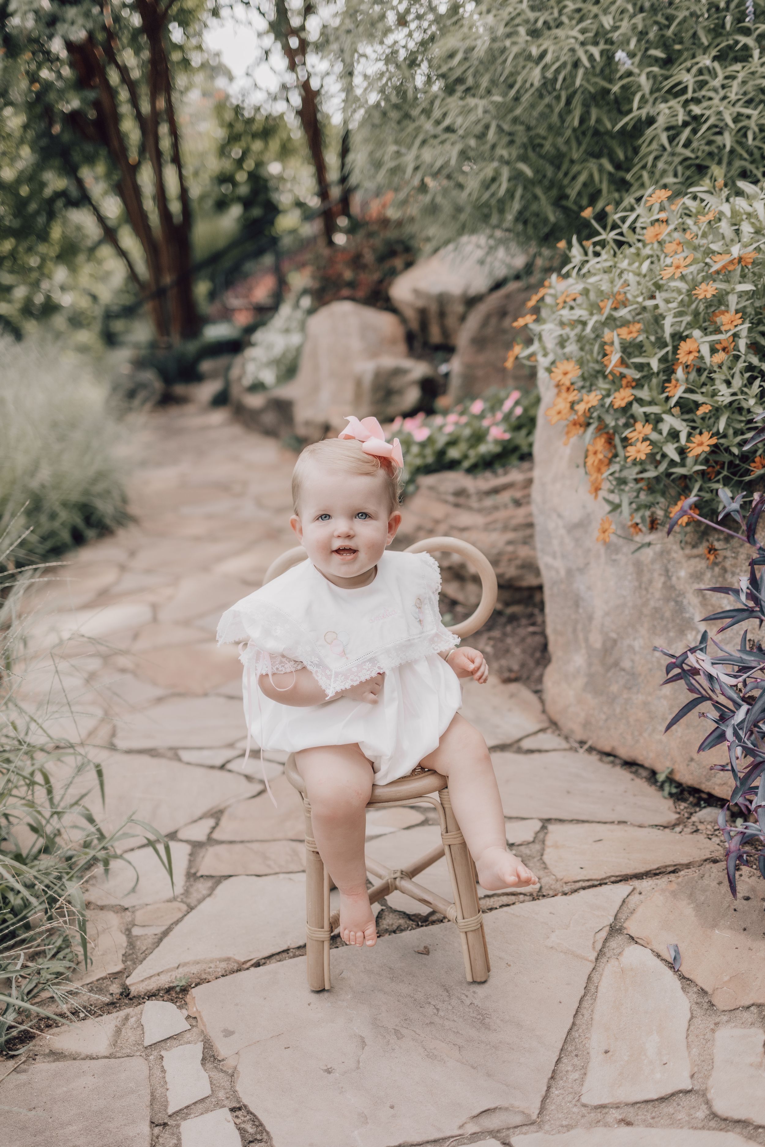 In a garden setting with stone paths and orange flowers, a baby sits in a small wooden chair wearing a white dress.