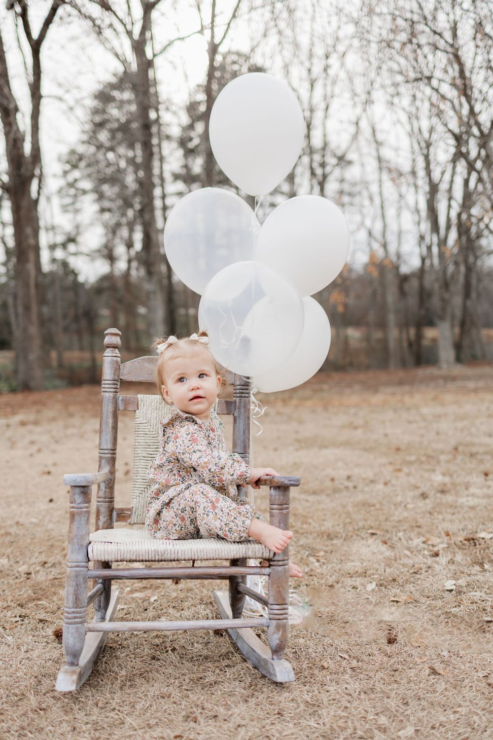 A child sits in a gray rocking chair with white balloons in an autumn outdoor setting.