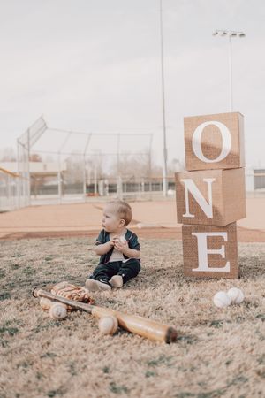 Rustic baseball-themed first birthday photo with wooden blocks spelling ONE beside a small bat and baseballs on a field.