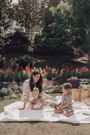 A family enjoys quality time together while lounging on a white picnic blanket in a garden with blooming flowers.
