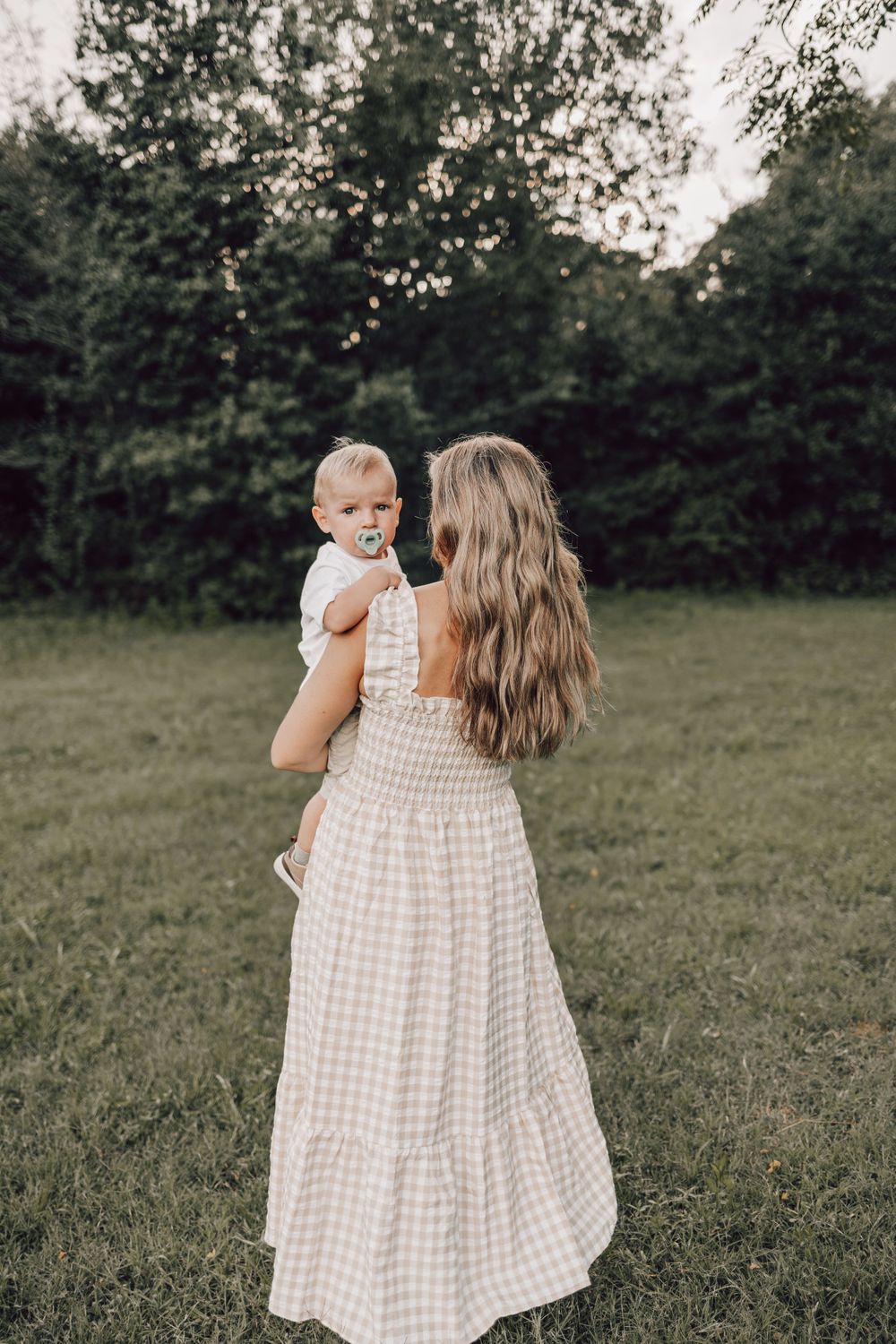 A person in a gingham dress holds a baby while standing in a grassy field at dusk.