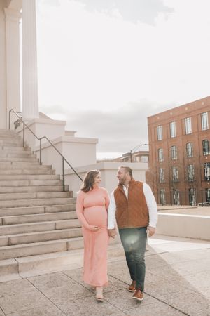 A couple in pink dress and brown jacket hold hands while walking on concrete steps outdoors on an overcast day.
