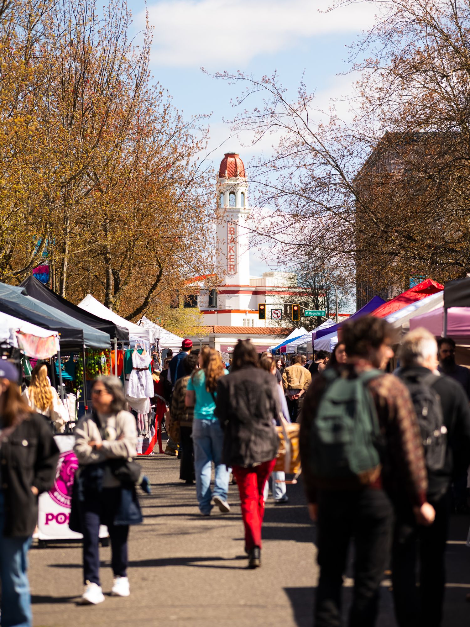 Busy outdoor market with clock tower visible through autumn trees.