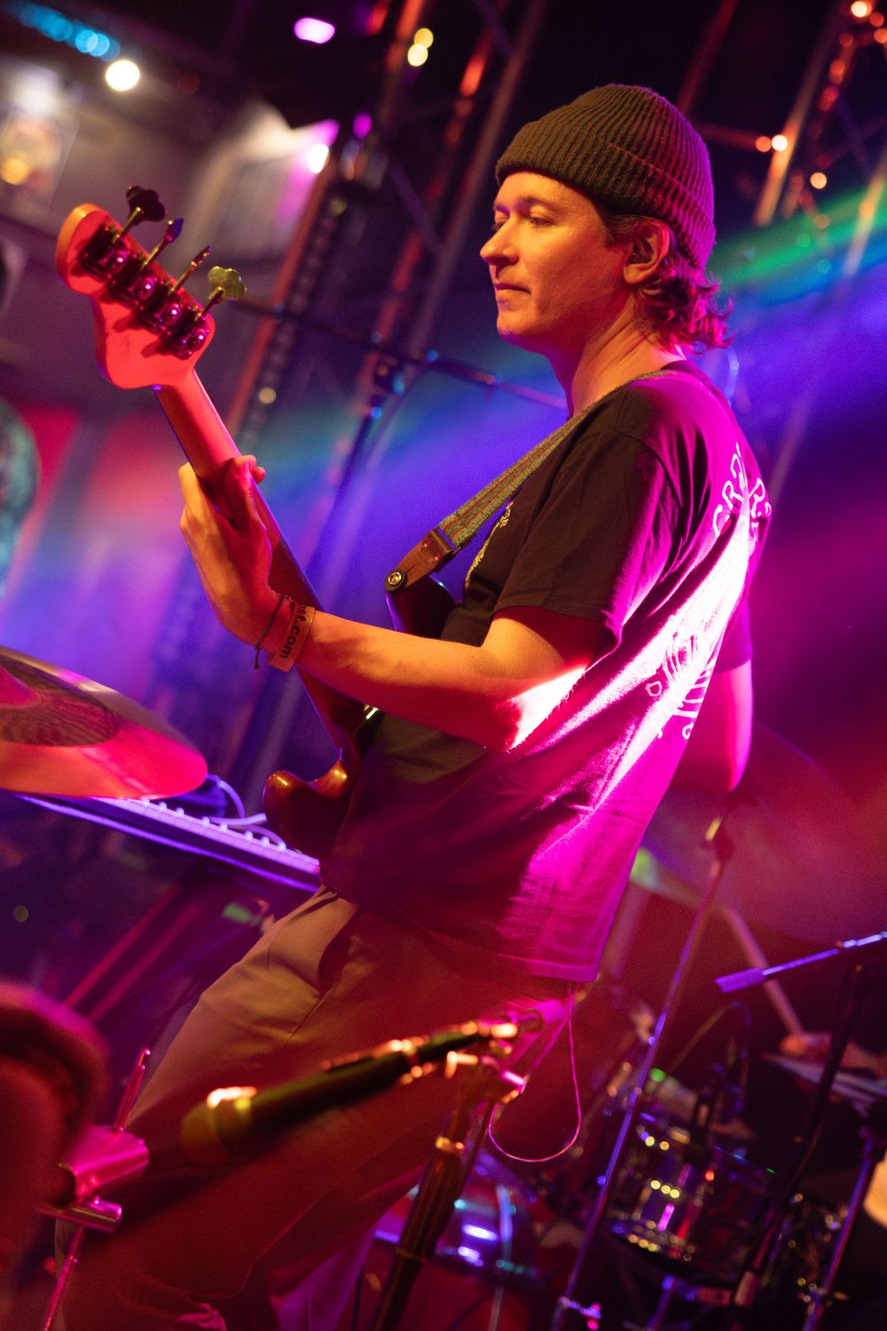 Bass guitarist performs under colorful stage lights wearing a beanie and t-shirt.