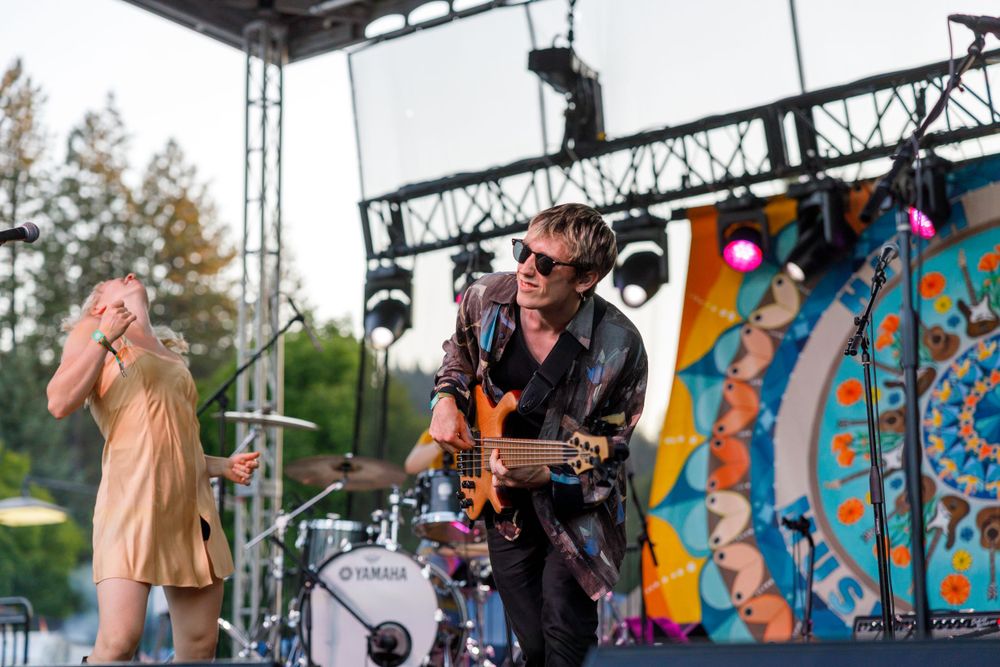 Musicians perform on an outdoor festival stage with colorful psychedelic backdrop and professional lighting setup.