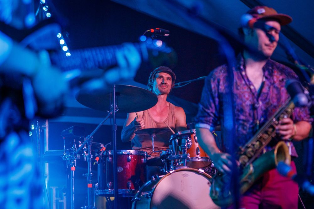 Drummer plays red drum kit with saxophone player in blue-tinted stage lighting.