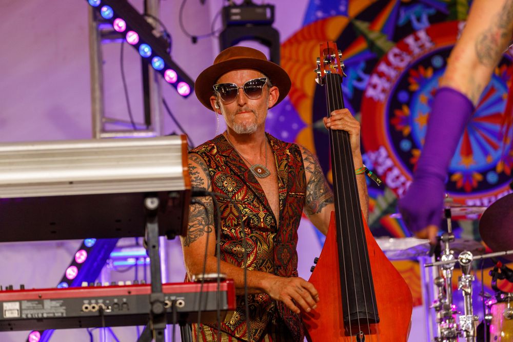 A musician plays bass while performing in front of a colorful psychedelic backdrop.