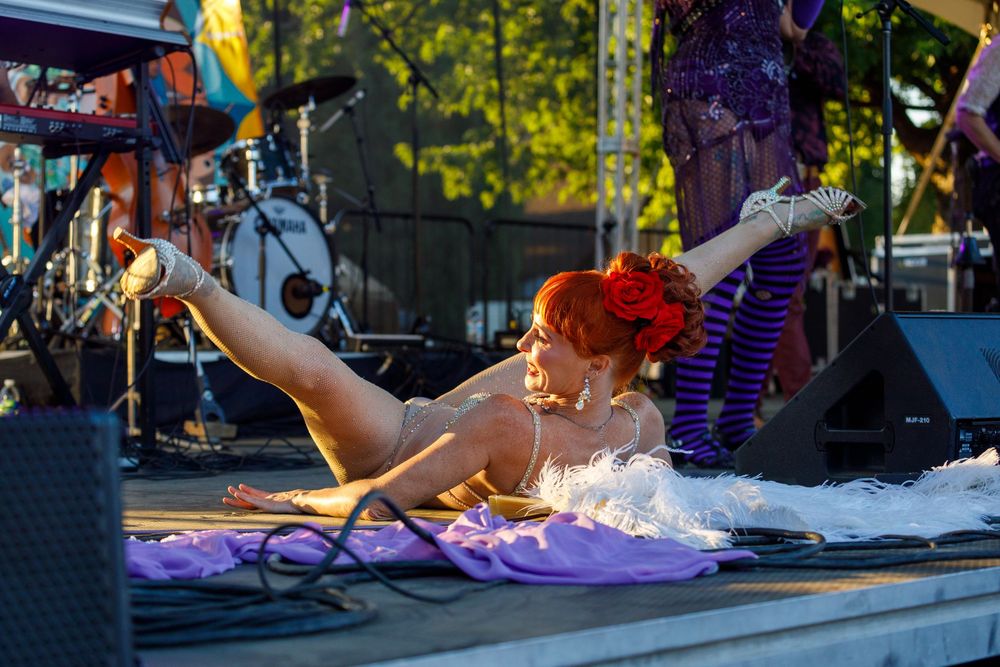 A performer in a white tutu and red flower lies on a colorful stage during an outdoor burlesque performance.