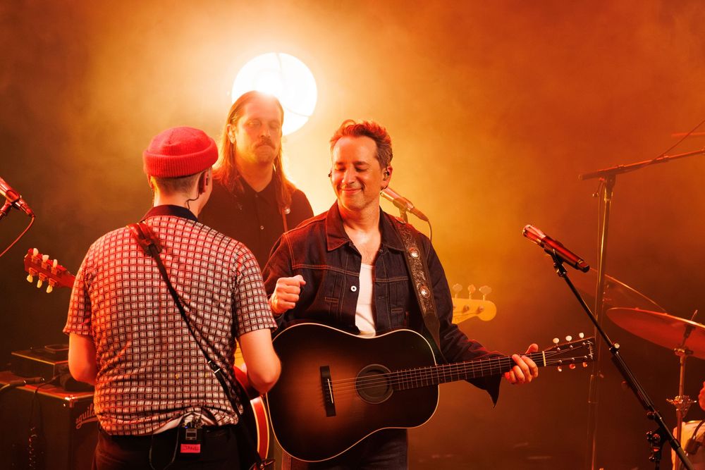 Musicians perform on stage with dramatic orange lighting and smoke effects during a live concert performance.