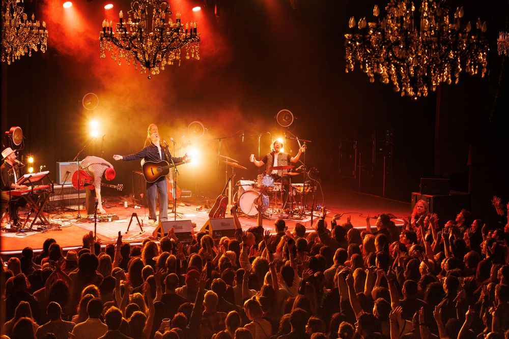 Concert performers on stage bathed in dramatic red and orange lighting while an energetic crowd watches from below.