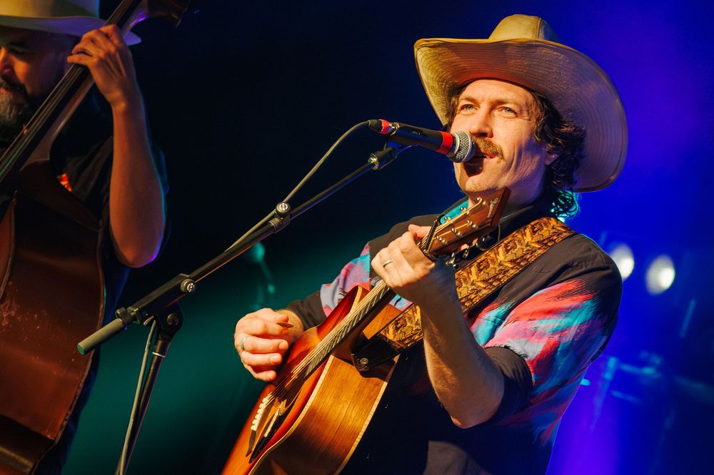 Country musician performs on stage with acoustic guitar under blue stage lighting during live concert performance.