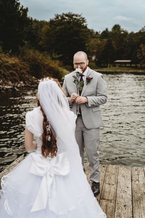 Bride and groom share a romantic moment on a wooden dock by a lake, with groom smelling a white rose.