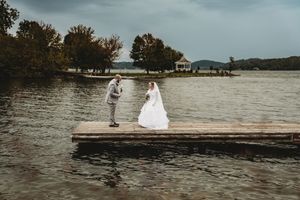 Bride and groom standing on a wooden dock over a lake surrounded by trees on their wedding day.