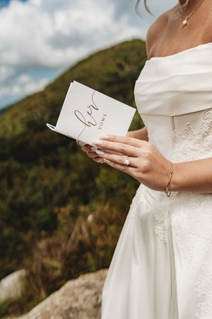 Bride in white off-shoulder gown holding her vows booklet outdoors with green hills in the background.