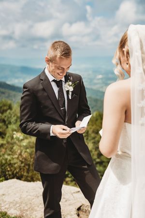 Groom in black suit reads wedding vows from card while smiling, standing outdoors with mountain landscape backdrop.