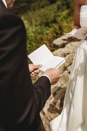 Groom in black suit reads wedding vows from a card during outdoor ceremony, bride in white dress visible.