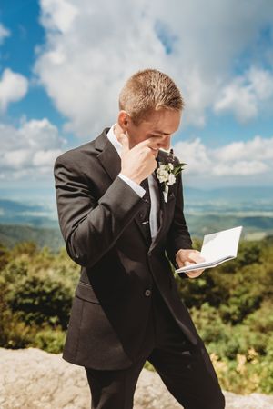 Emotional groom in black suit reading wedding vows outdoors, wiping tears with scenic mountain landscape background.
