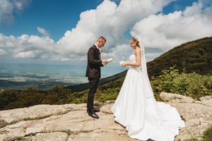 Couple exchanging vows on a rocky mountain summit, bride in white gown, groom in black suit, scenic valley backdrop.
