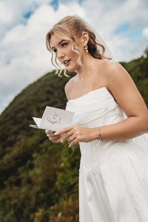 Blonde bride in strapless white gown holding vows card outdoors with green hills and cloudy sky backdrop.
