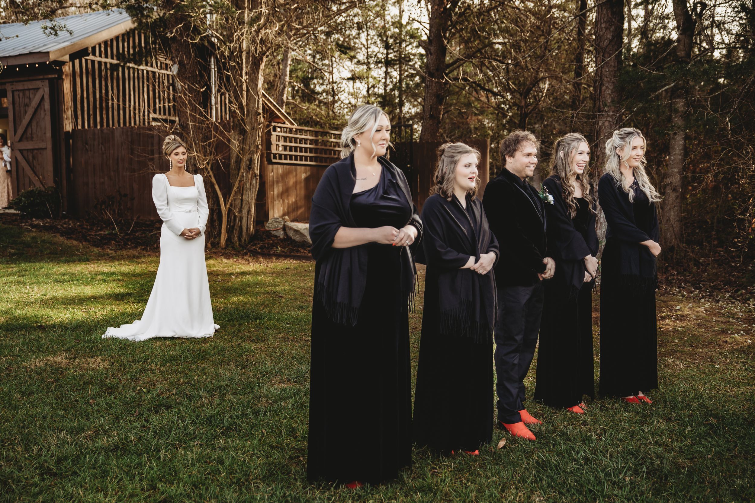 Wedding party in black dresses stands in line while bride poses separately.