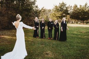 Bride in white dress gestures to bridesmaids in black dresses on lawn.