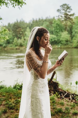 Tattooed bride in lace wedding dress and veil reads vows by a serene forest lake on her wedding day.
